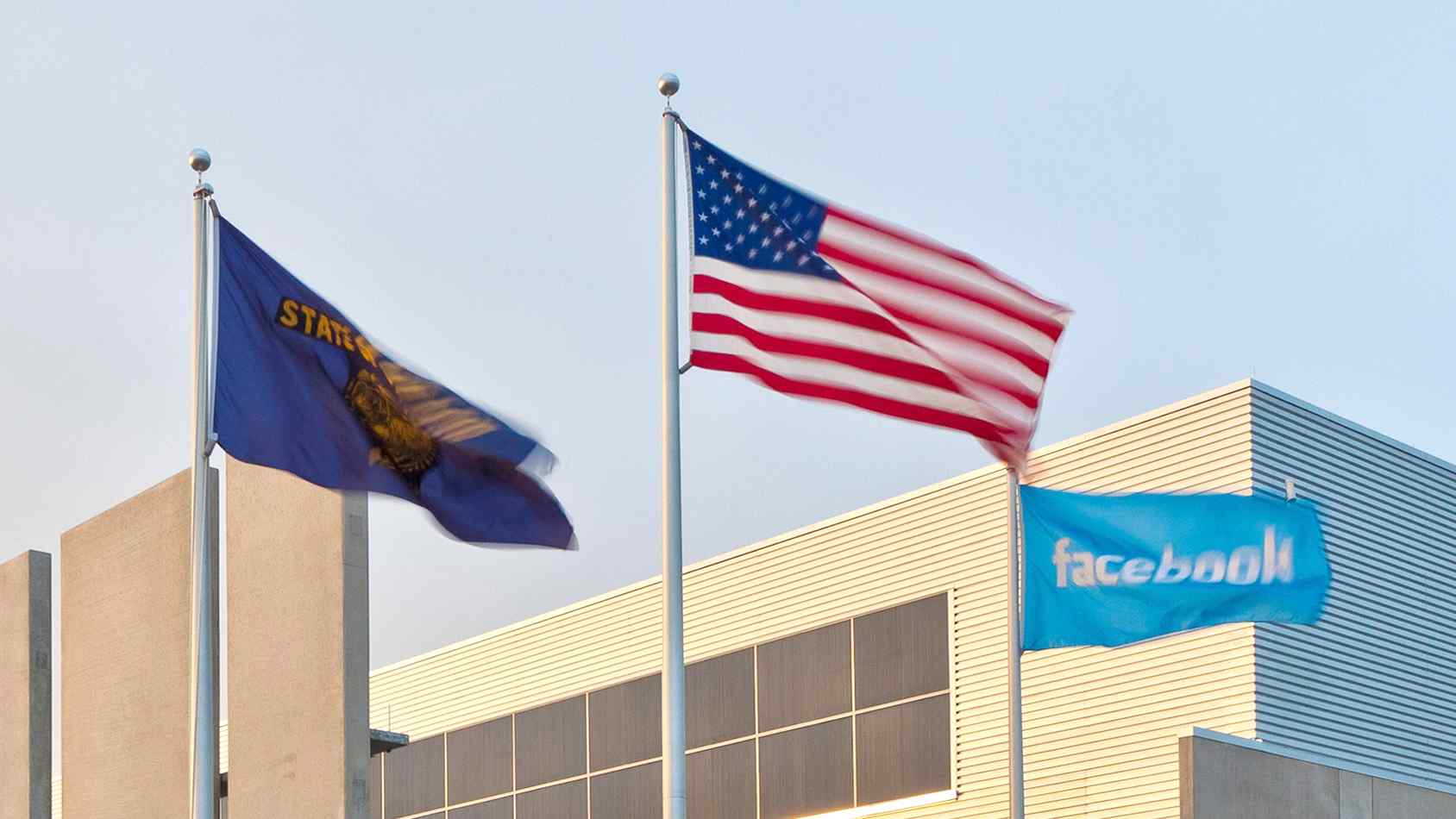 Three flags wave in the wind: the Oregon state flag, the American flag, and a Facebook flag, set against a modern building backdrop.