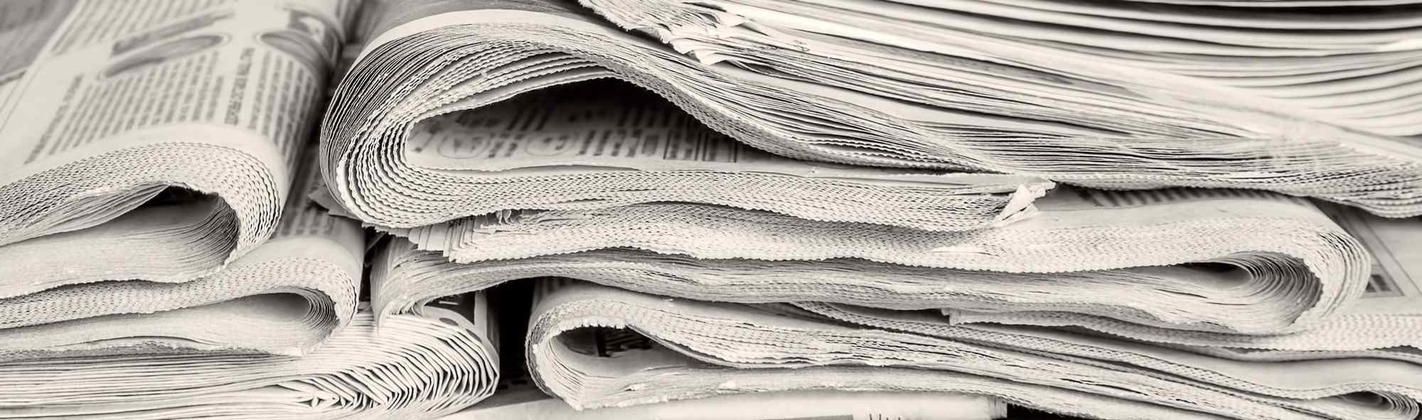 A close-up of stacked newspapers, showcasing their folded edges and textured pages in black and white.