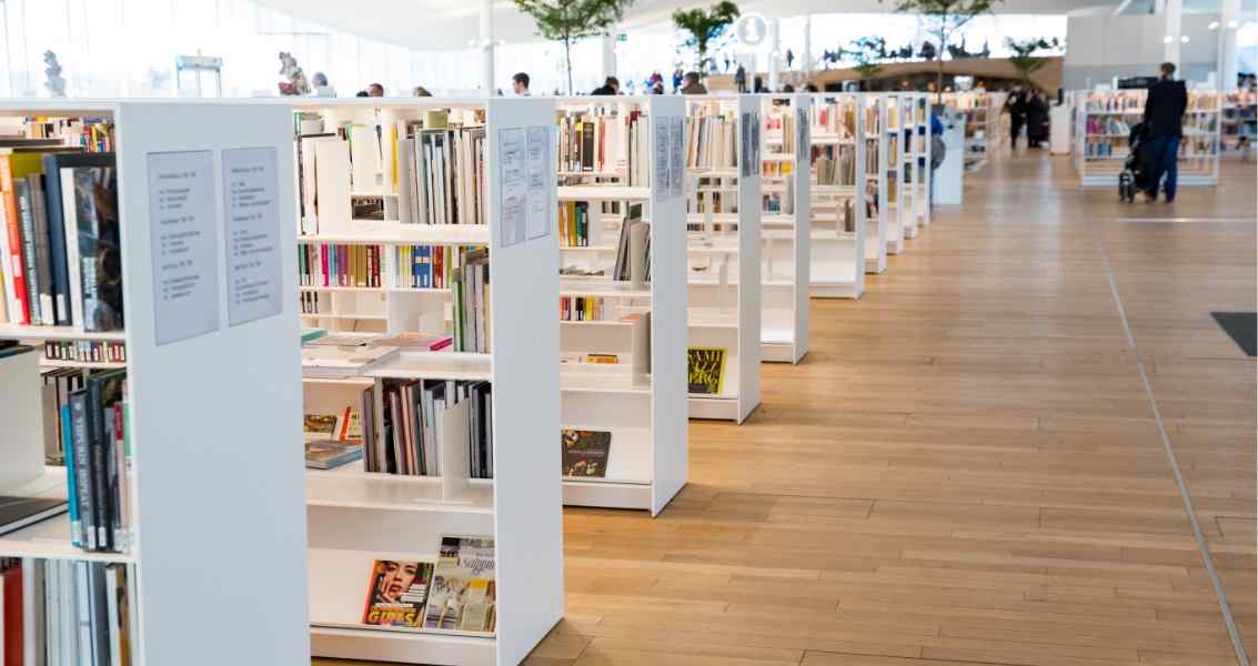 A modern library interior featuring angular white bookshelves filled with various books, surrounded by people browsing and reading.