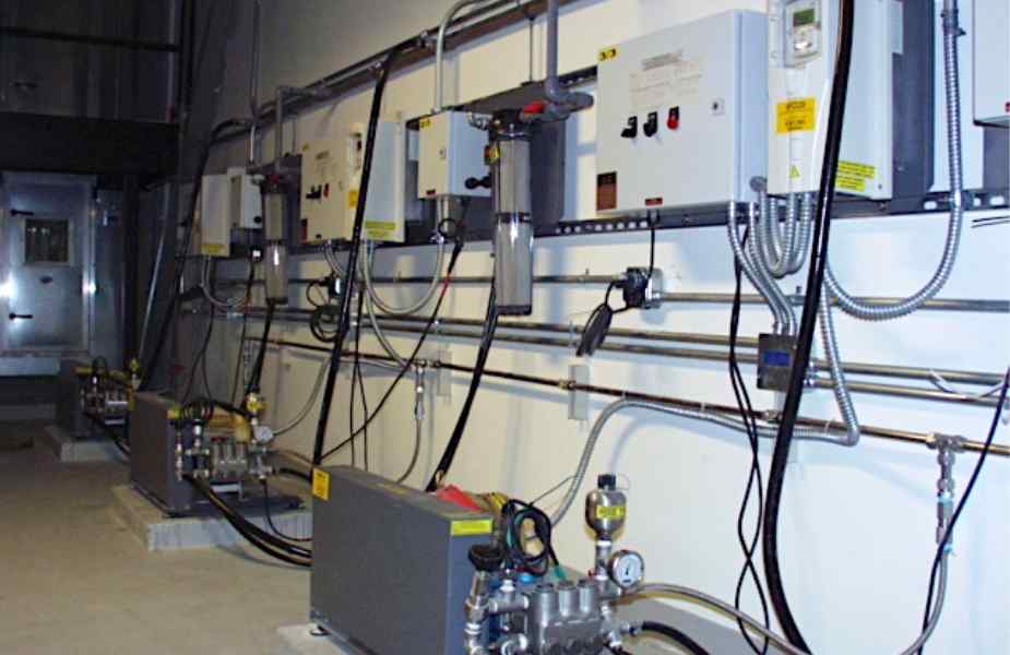 View of industrial equipment with control panels and piping along a wall, featuring multiple pumps and electrical connections.