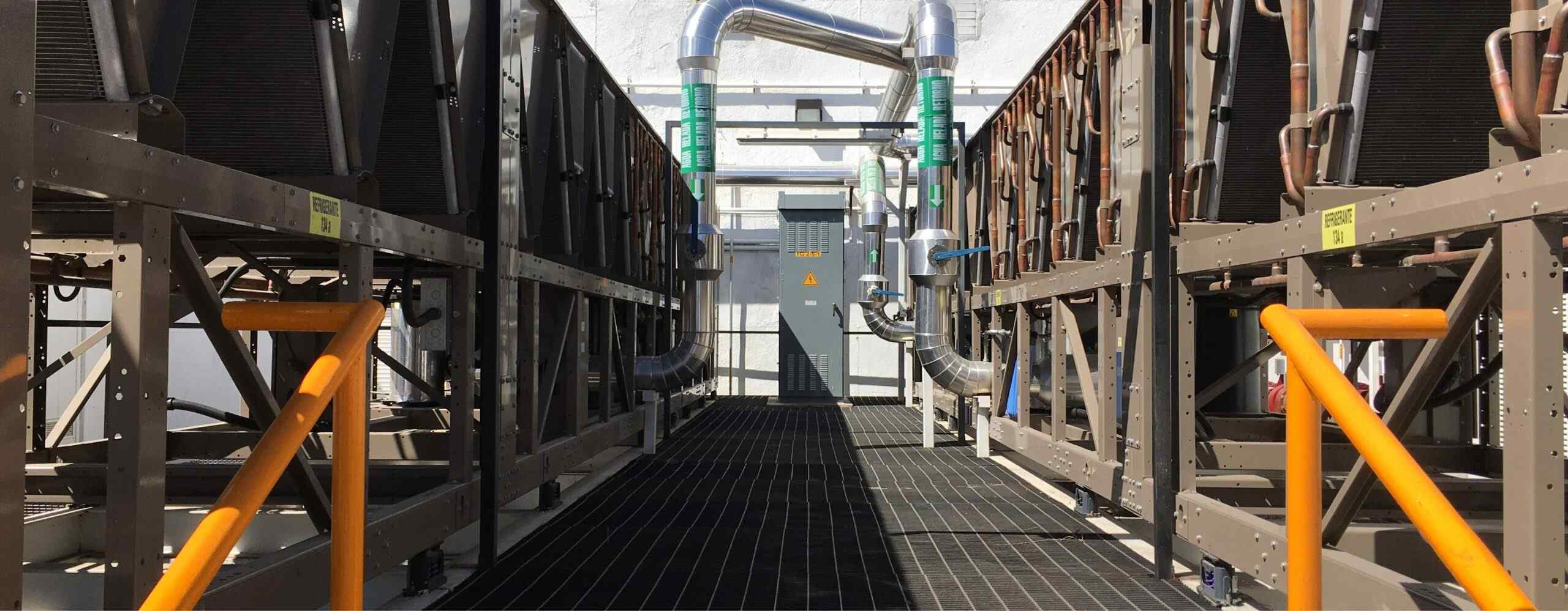 Industrial HVAC system with large cooling units, metal pipes, and an orange safety railing under bright sunlight.