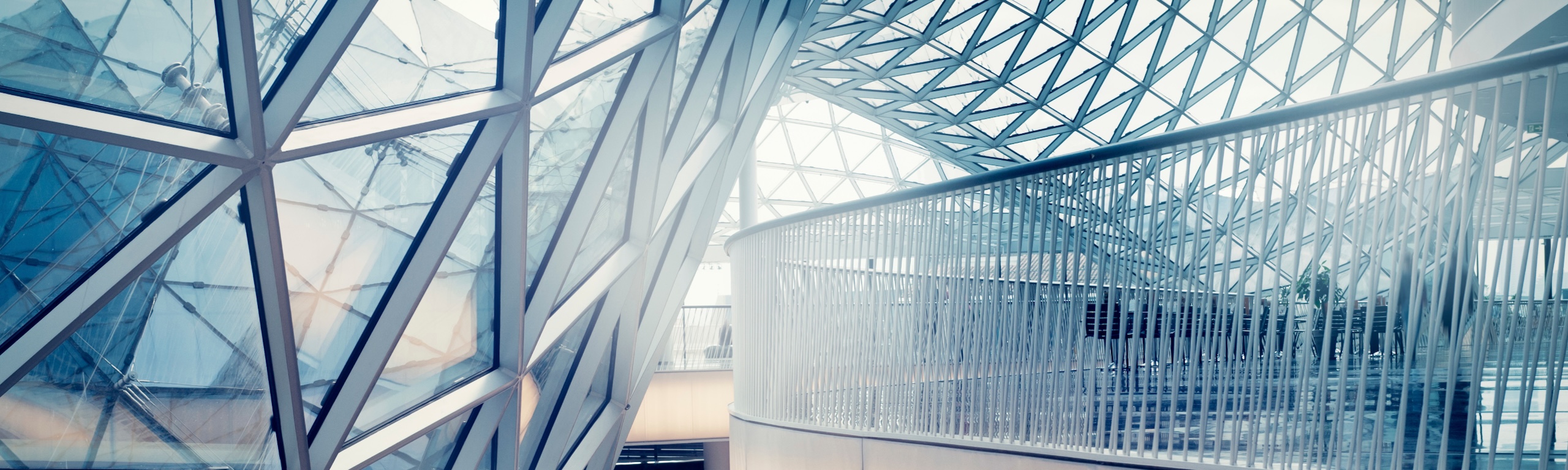 Interior of a futuristic building featuring a curved walkway enclosed by a sweeping geometric glass dome and walls with a distinct triangular lattice framework.