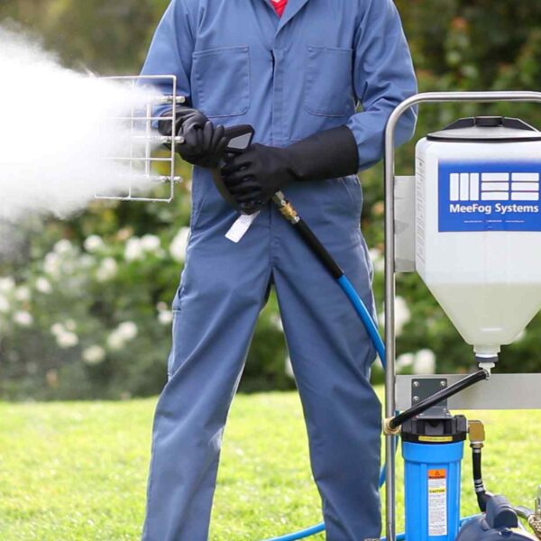 A person in a blue jumpsuit and gloves operates a fogging machine, spraying mist over grassy greenery. The machine brands "MeeFog Systems."