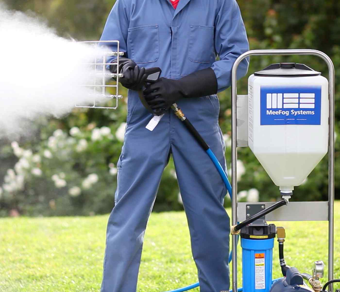 A person in a blue jumpsuit and gloves operates a fogging machine, spraying mist over grassy greenery. The machine brands "MeeFog Systems."
