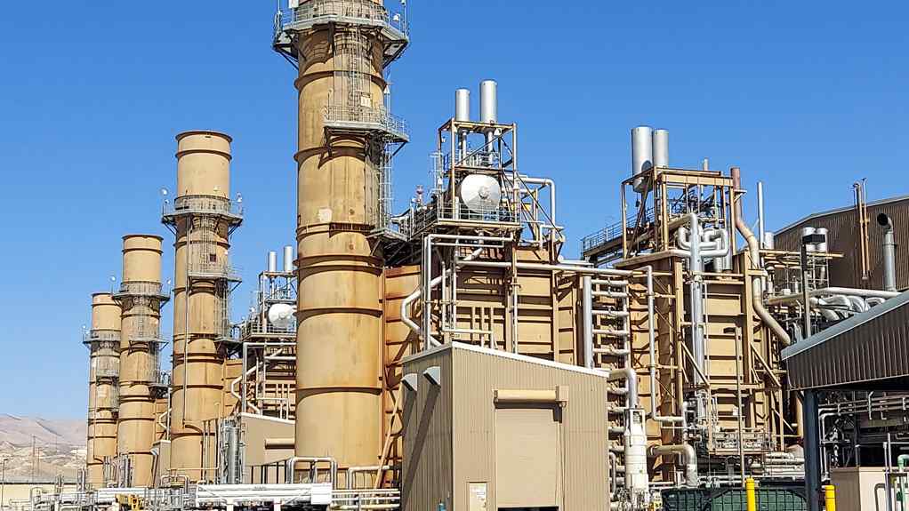 A large industrial facility featuring several tall smokestacks, pipelines, and equipment against a clear blue sky.