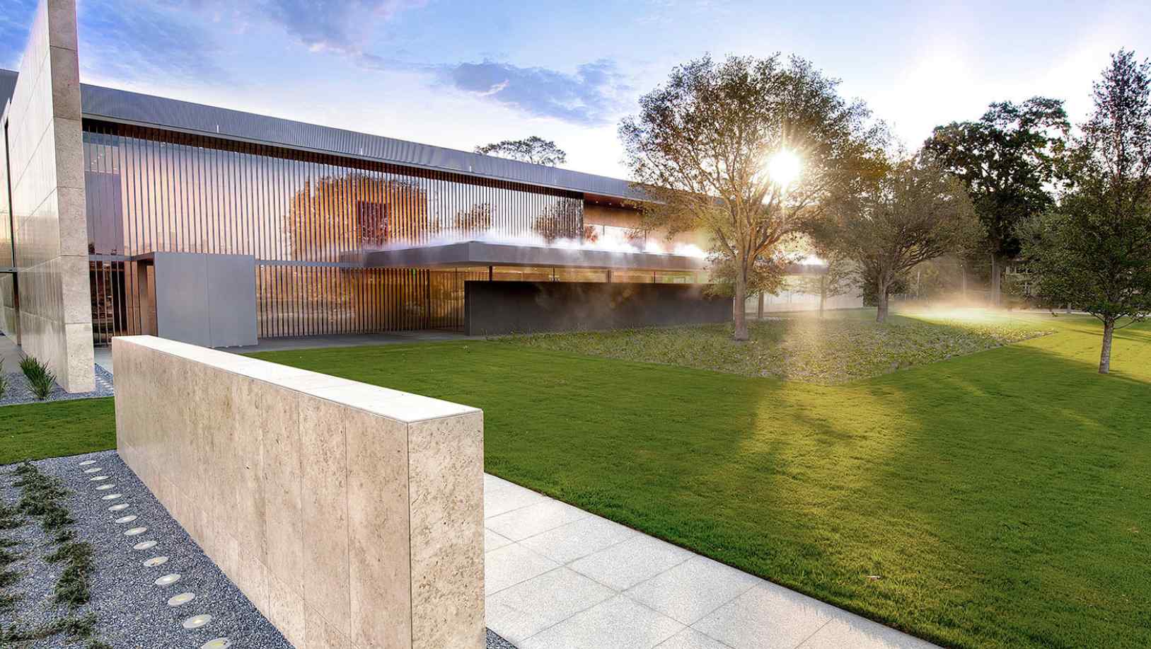 Modernist building with reflective metal facade, surrounded by lush green lawn and trees, illuminated by a warm, morning sun.