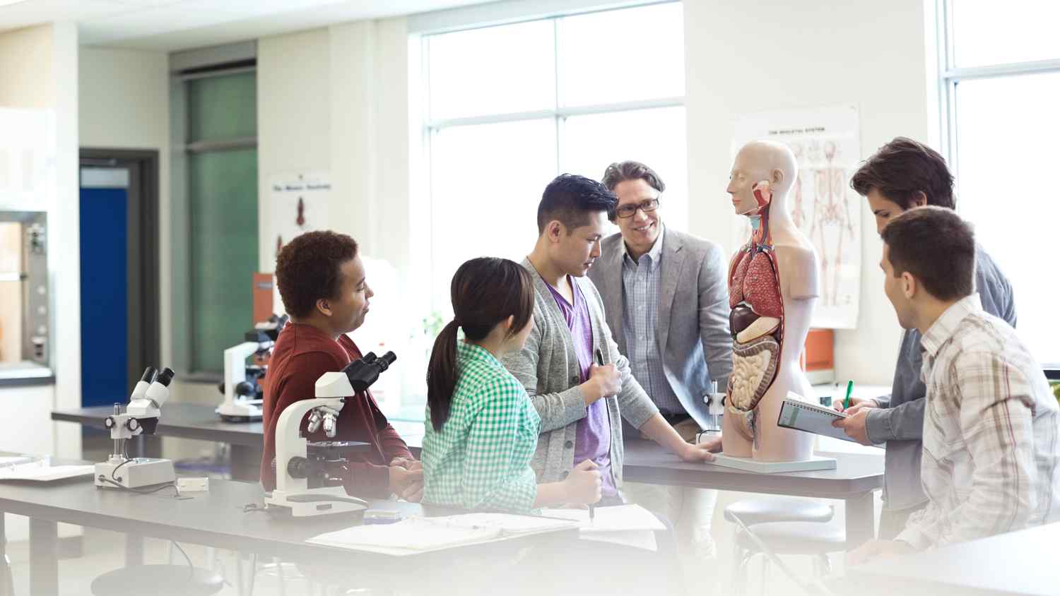 A diverse group of students observe a life-sized anatomical model in a bright laboratory, with microscopes and notes on the table.