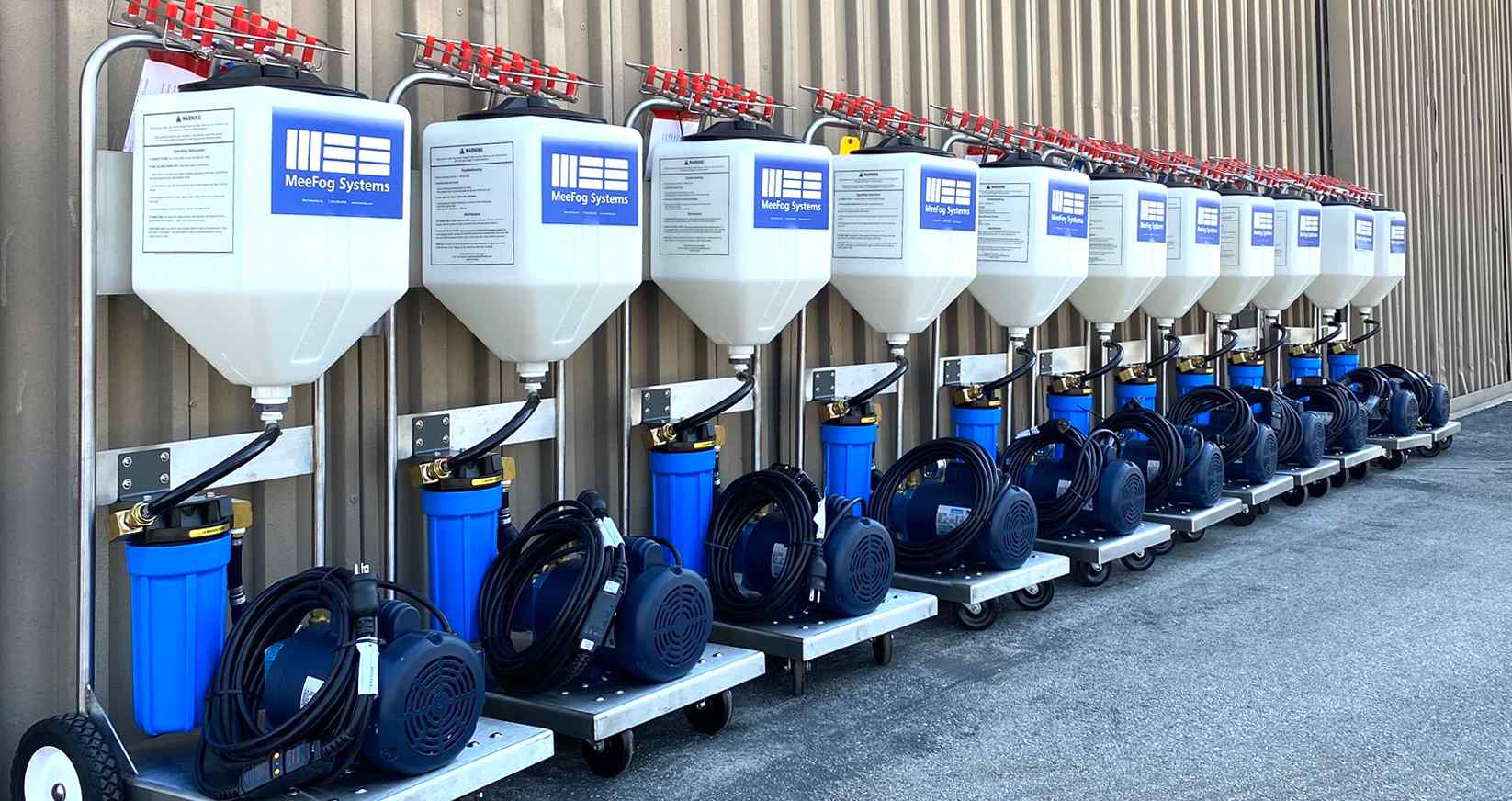 A row of ten MeeFog misting systems on wheeled carts, featuring tanks, filters, and electric motors, arranged against a metal wall.
