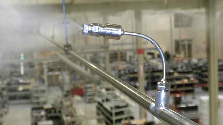 A stainless steel misting nozzle sprays water vapor from a metallic pipe in a factory filled with equipment and machinery.