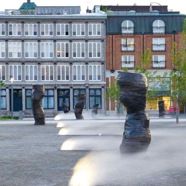 A modern plaza featuring abstract sculptures surrounded by mist, with historic buildings and lush trees in the background.