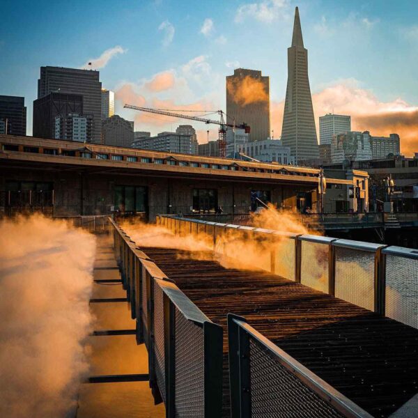 A wooden pier emits steam, framed by San Francisco's skyline and the Transamerica Pyramid under a colorful sunset.