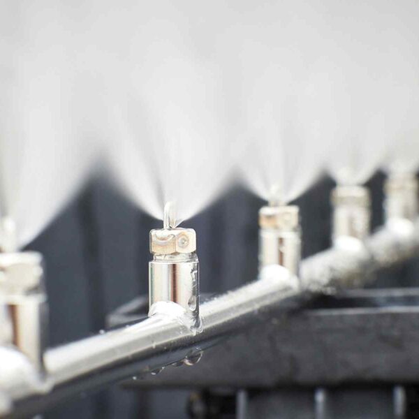 A close-up of metal misting nozzles spraying fine water droplets, creating a cooling effect in a bright, out-of-focus background.