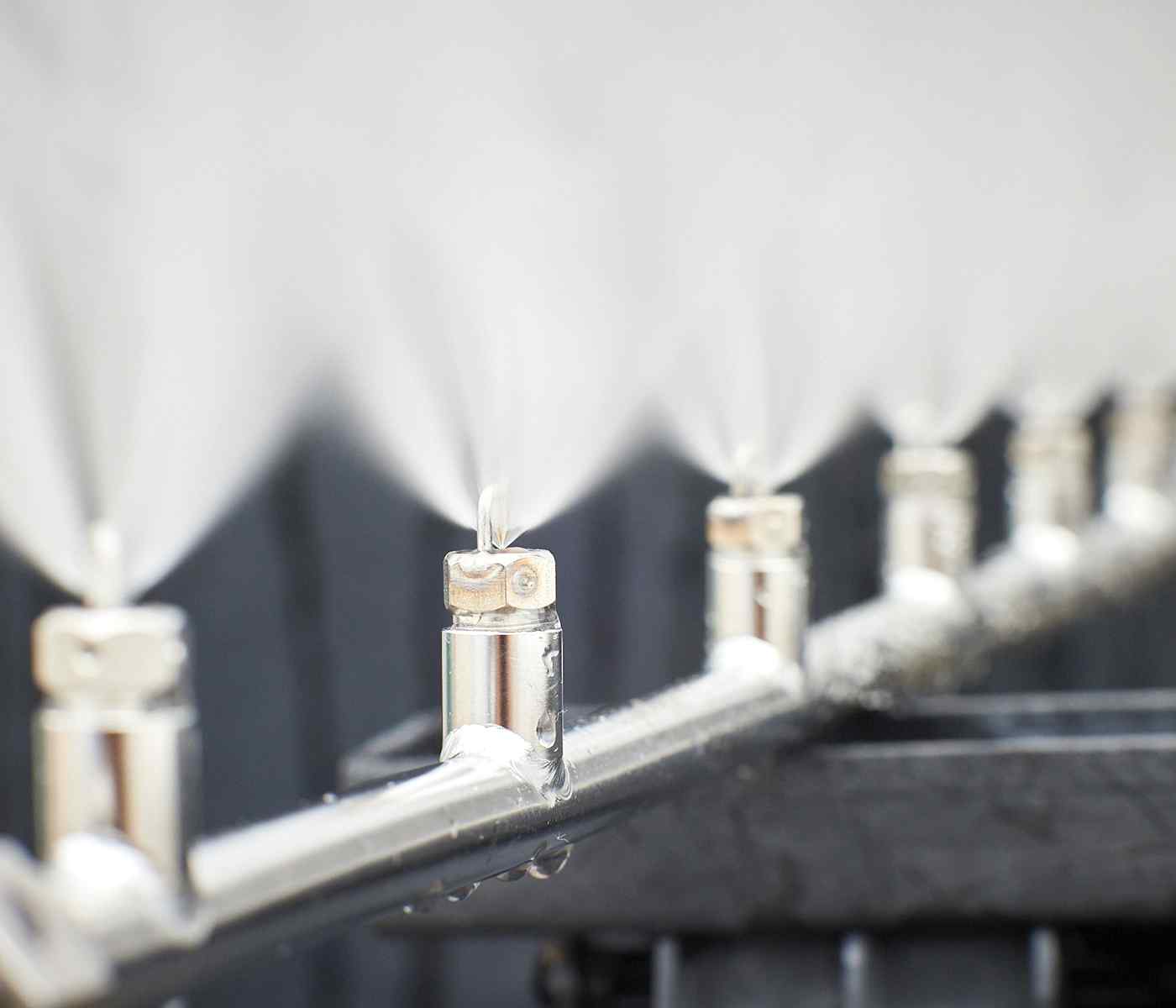 A close-up of metal misting nozzles spraying fine water droplets, creating a cooling effect in a bright, out-of-focus background.