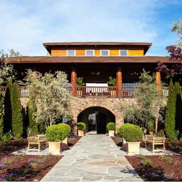 A beautiful stone and wood building with a wide entrance, flanked by lush gardens and vibrant trees under a clear blue sky.