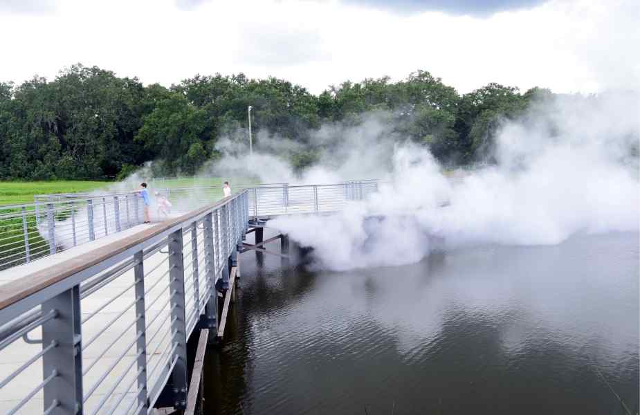 A misty scene on a walkway over water, with people walking amidst fog rising from the surface, surrounded by lush greenery.