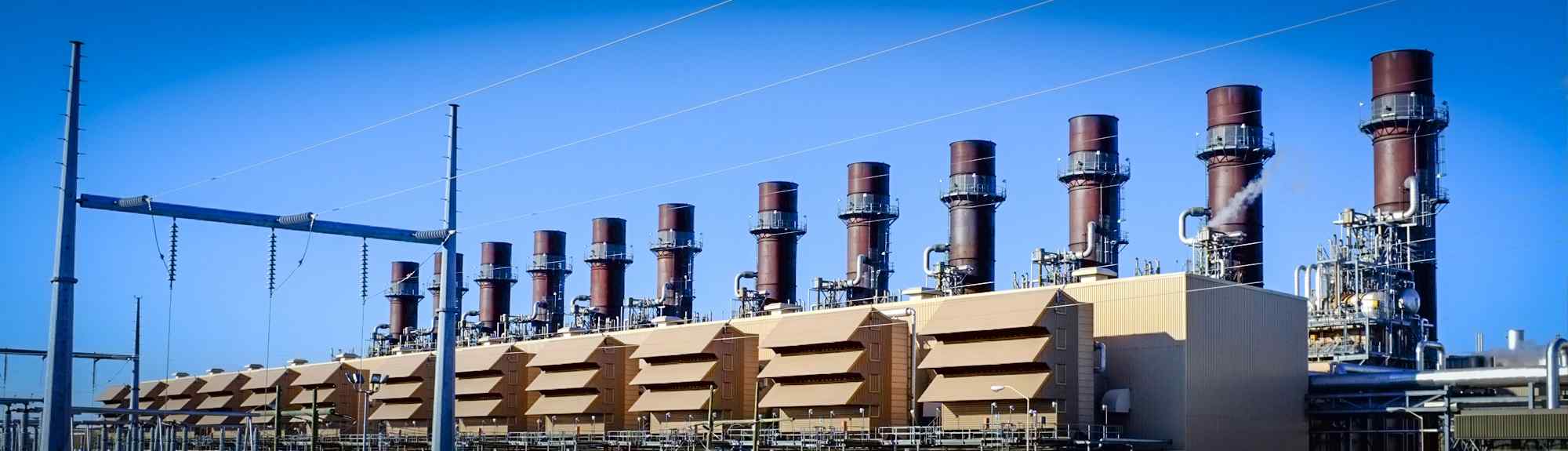 A gas-fired power plant with multiple tall exhaust stacks, featuring industrial structures, power lines, and a clear blue sky.