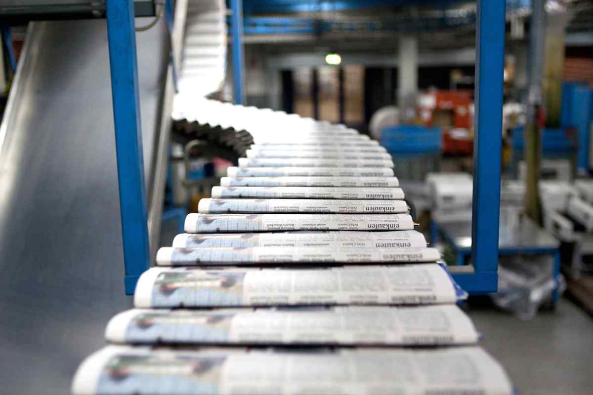 A close-up view of rolled newspapers on a conveyor belt in a printing facility, with blurred machinery and equipment in the background.