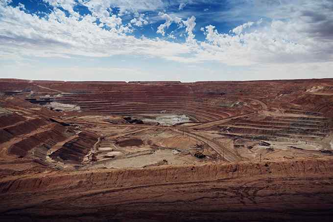 A vast open-pit mine with terraced layers, surrounded by rugged terrain under a cloudy blue sky.