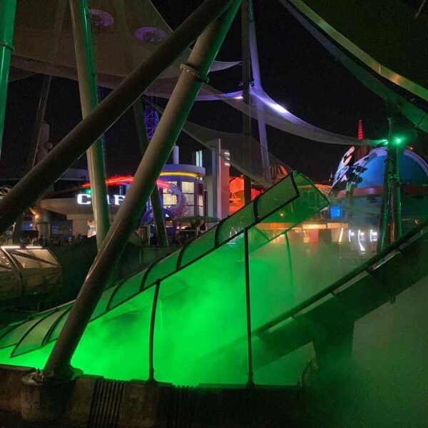 Nighttime view of a neon-lit amusement park slide with green mist at the base, surrounded by vibrant attractions and illuminated structures.