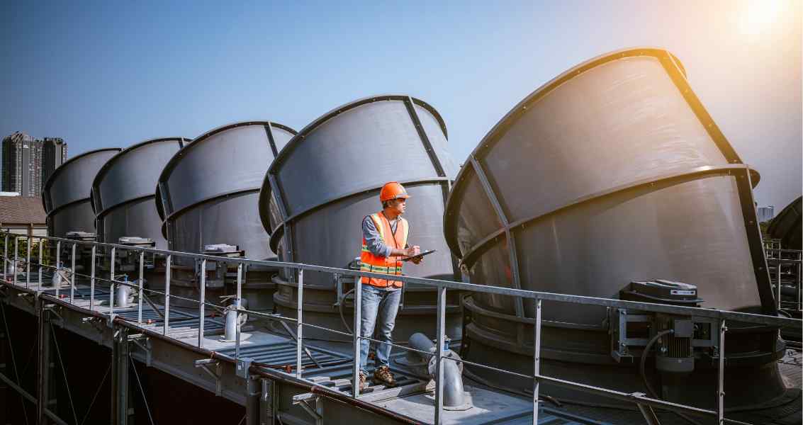 A worker in a hard hat and safety vest stands on a rooftop, checking equipment next to large cooling units against a bright sky.