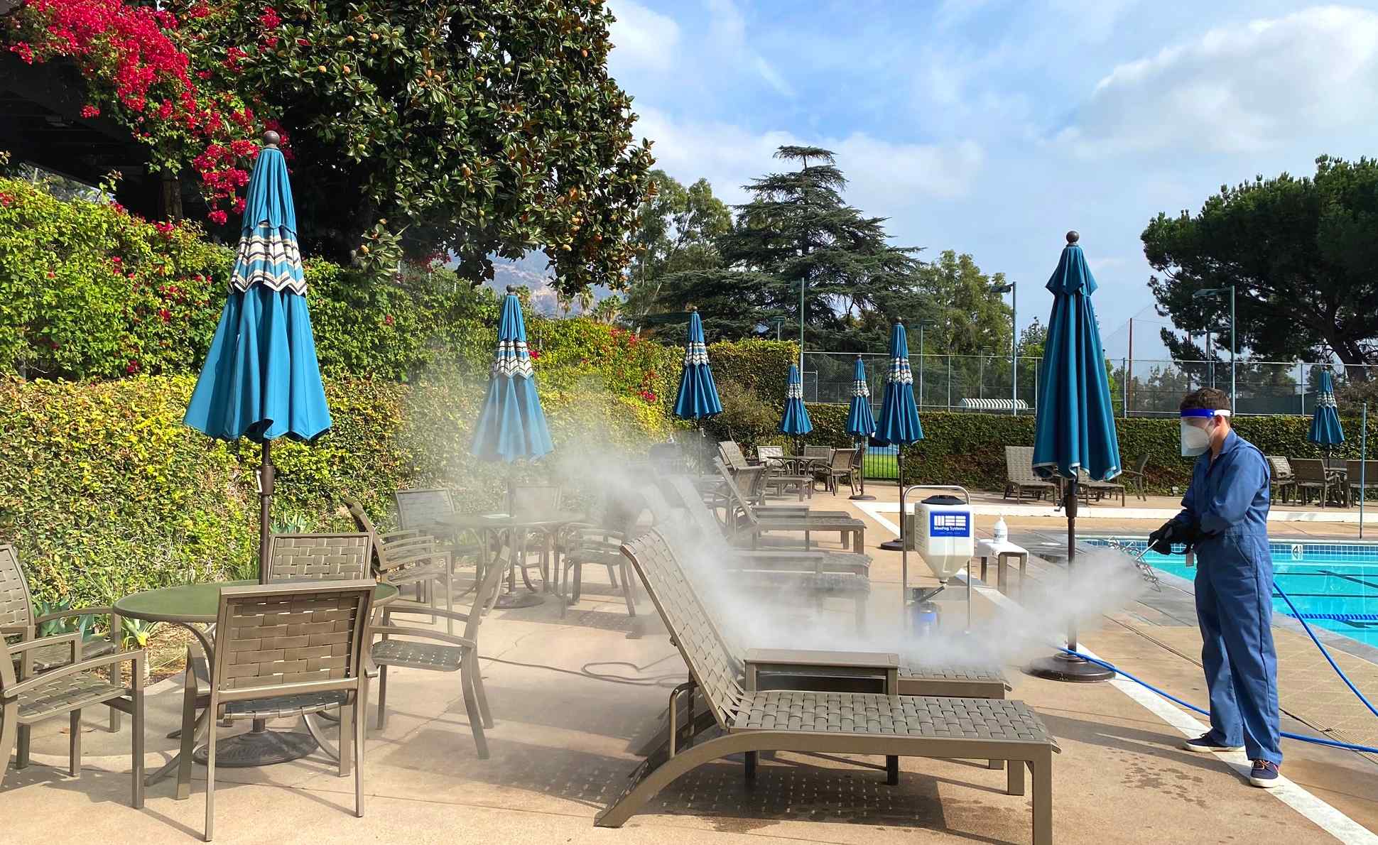 A worker in blue overalls sprays mist over poolside lounge chairs, surrounded by green shrubs and blue umbrellas, beside a swimming pool.
