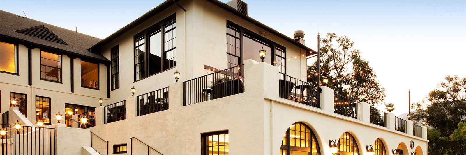 A beautiful two-story building features large windows, a balcony with lights, and a charming terrace, set against a clear blue sky.