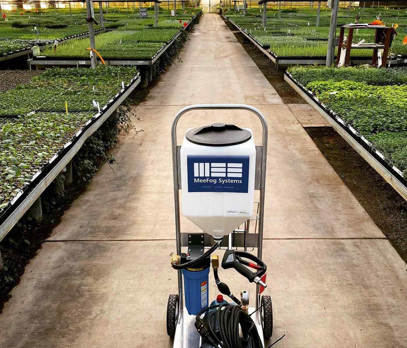 A fogging machine with a MeeFog Systems logo sits on a path surrounded by rows of vibrant, growing plants in a greenhouse.