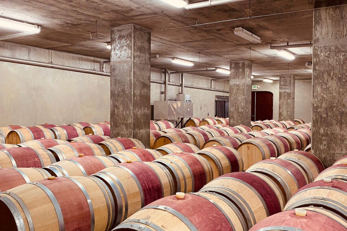 Rows of wooden wine barrels with red center stains resting on a concrete floor in a large cellar.