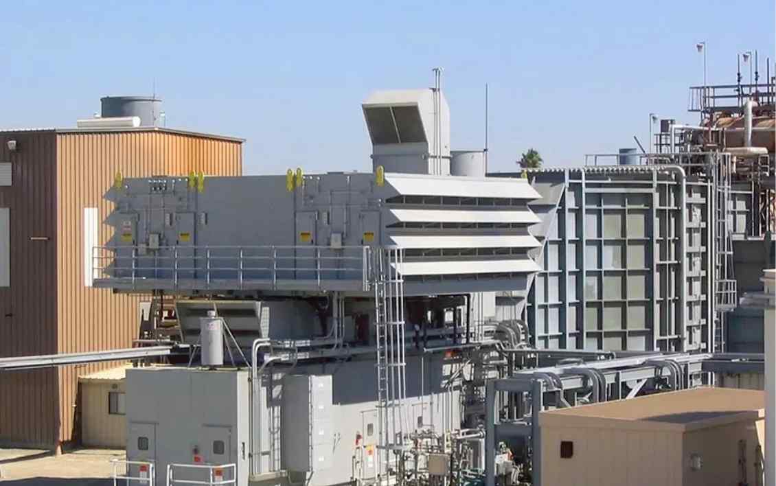 Industrial facility featuring large gray equipment, piping, and storage buildings under a clear blue sky.