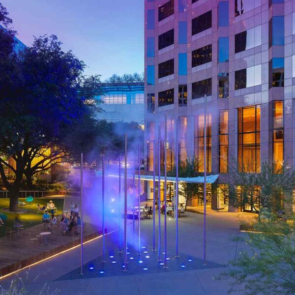 A vibrant outdoor plaza at dusk featuring a colorful water feature, surrounded by people relaxing and modern buildings.