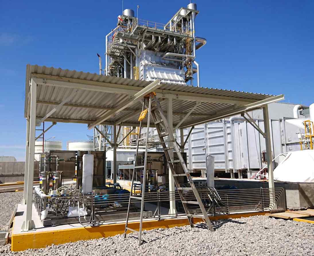 Industrial facility with machinery under a canopy, a ladder propped against it, and piping structures on gravel ground.