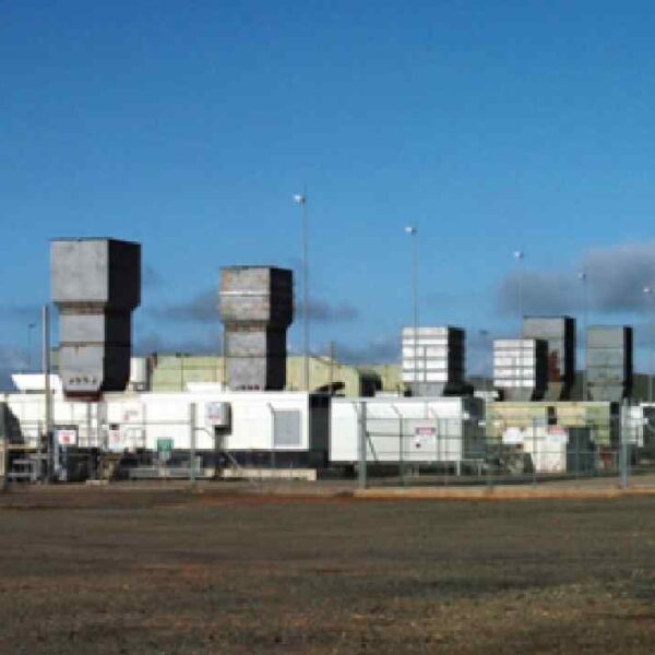 Industrial facility featuring large metal structures, storage containers, and equipment under a clear blue sky, enclosed by a fence.