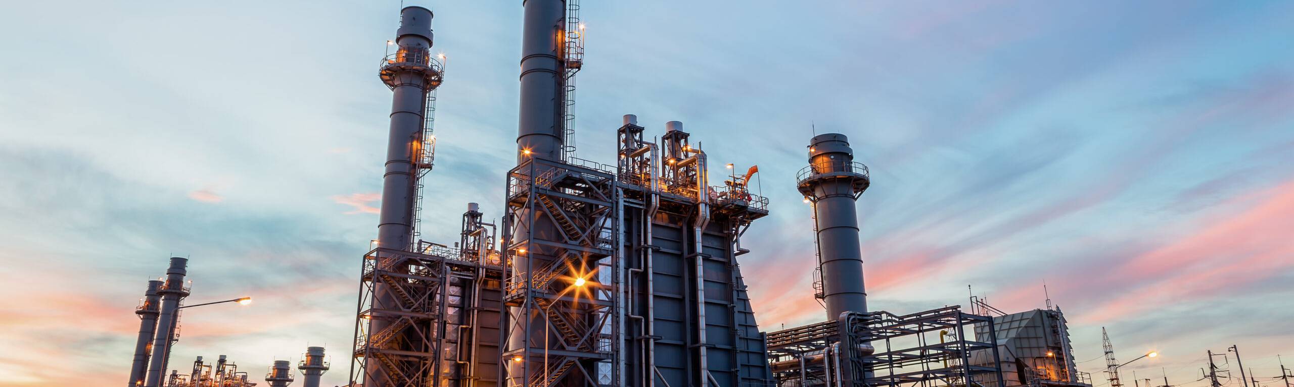 Gas-fired power plant at sunset, featuring tall smoke stacks, illuminated structures, and a vibrant sky.