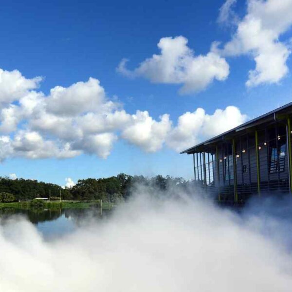 A modern building beside a serene water body, surrounded by fog and trees, under a bright blue sky with fluffy clouds.