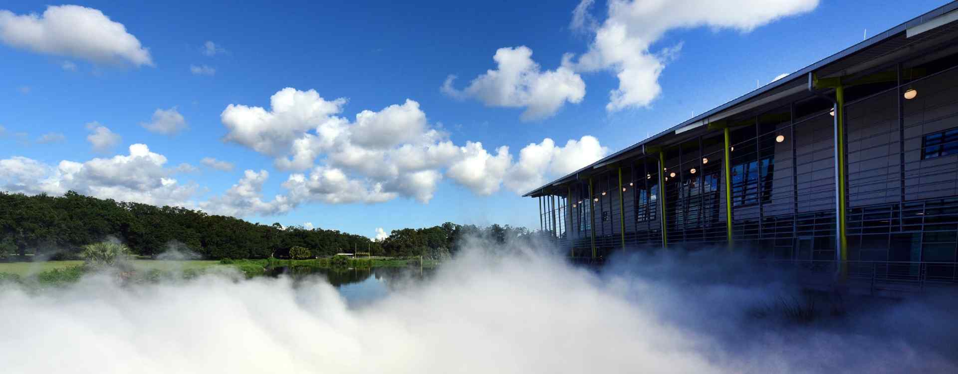 A modern building beside a serene water body, surrounded by fog and trees, under a bright blue sky with fluffy clouds.