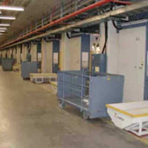 A spacious industrial hall featuring multiple blue machines and carts, with overhead pipes and bright lighting along a concrete floor.