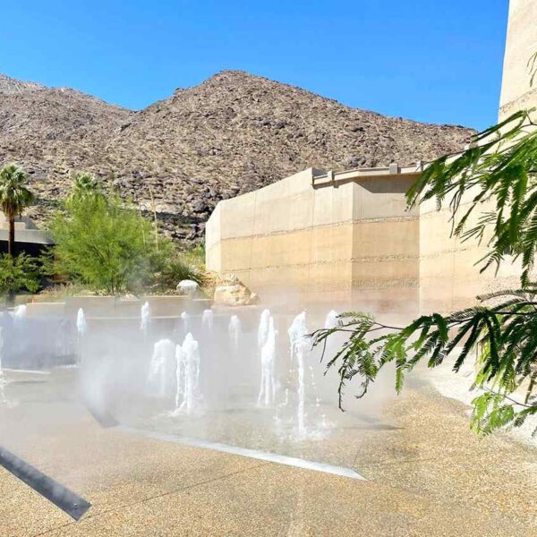 A tranquil scene with water fountains spraying in a misty plaza, surrounded by desert landscaping and distant mountains under a clear blue sky.