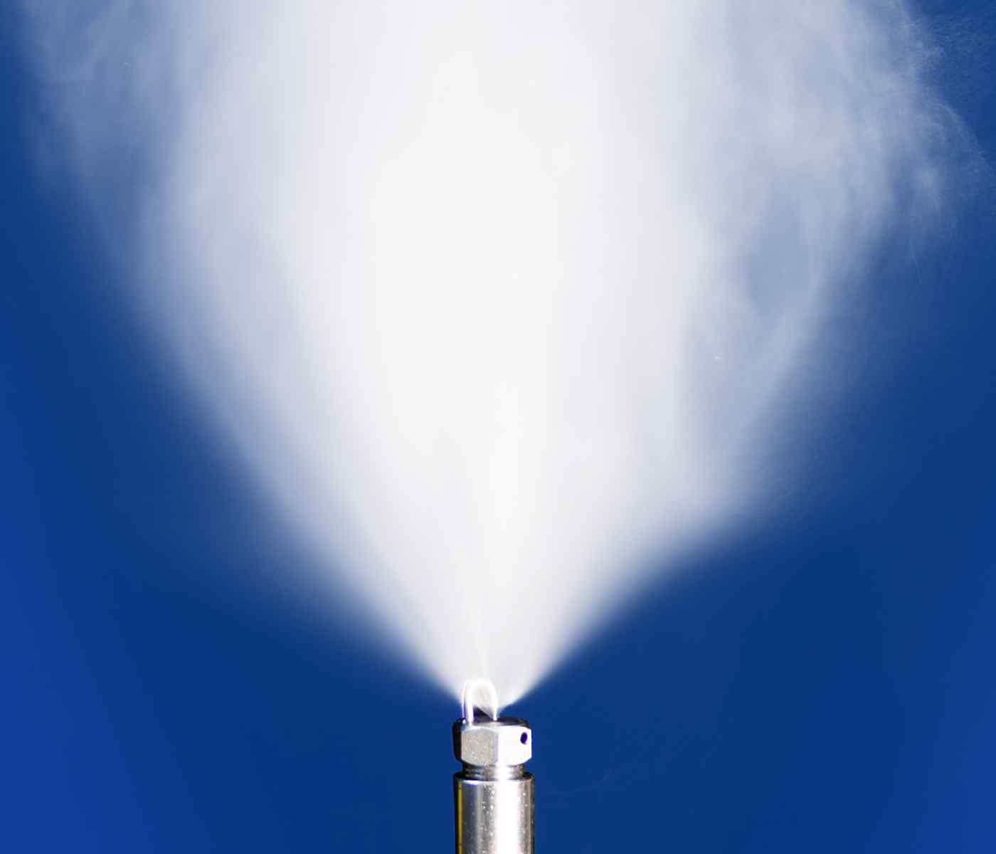 Close-up of a metallic nozzle emitting a fine mist against a blue background, creating a soft, white spray effect.