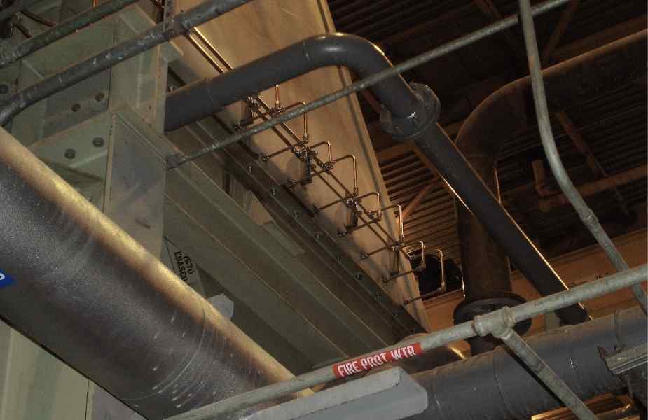 Pipes and industrial fittings overhead in a facility, showing a mix of metal structures and safety signage labeled 'FIRE PROT WTR.'