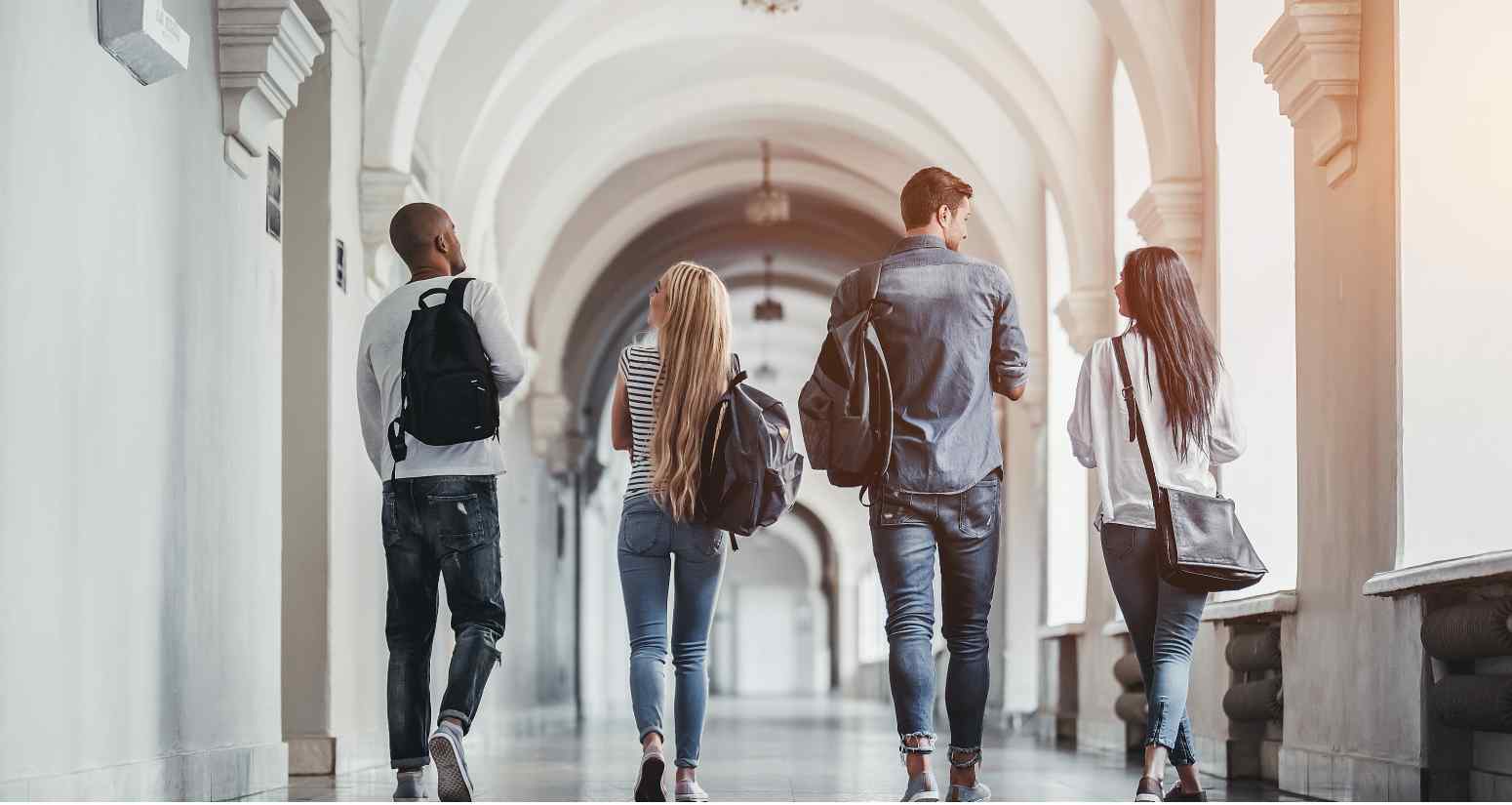 Four students walk down a long, arched corridor, each carrying backpacks, engaged in conversation, with bright light flooding in.