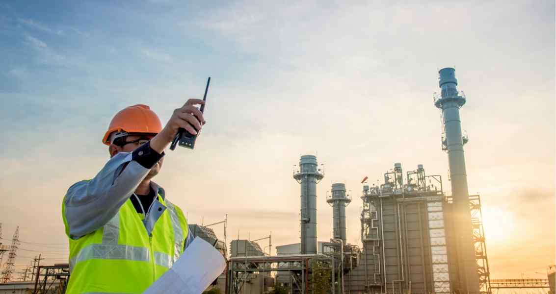 A worker in a hard hat and reflective vest speaks into a radio, with a power plant and smokestacks in the background at sunset.