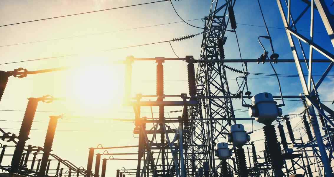 Silhouette of power lines and an electrical substation against a bright sunset sky, highlighting energy infrastructure.