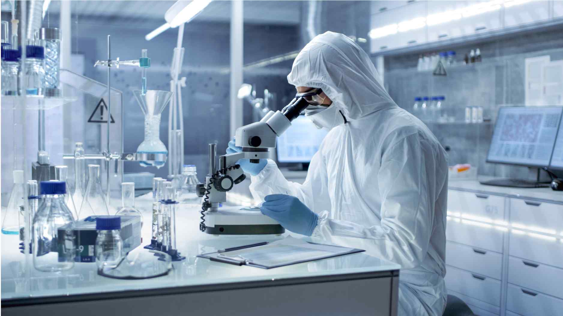 A researcher in a protective suit examines a sample under a microscope in a modern laboratory filled with glassware and equipment.