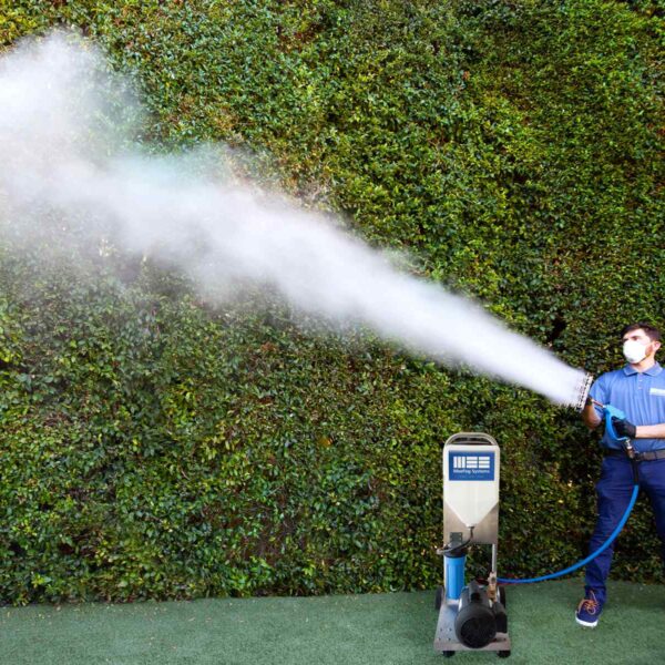 A person in blue attire operates a fog machine, releasing a thick stream of mist against a lush green backdrop.