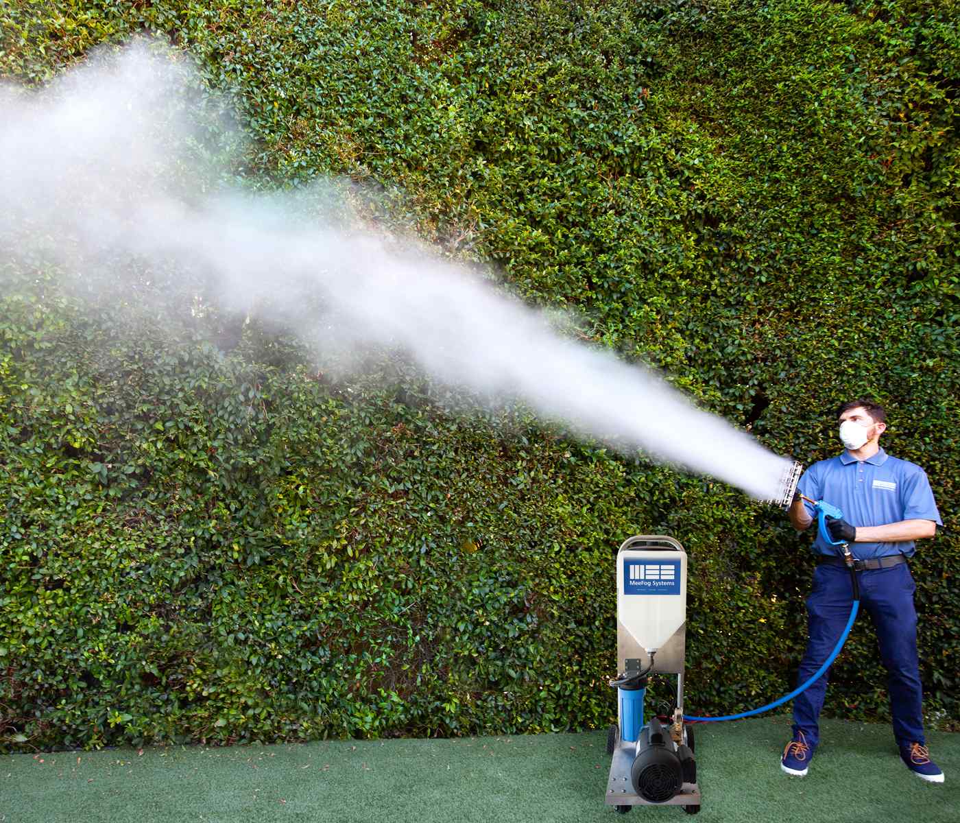 A person in blue attire operates a fog machine, releasing a thick stream of mist against a lush green backdrop.