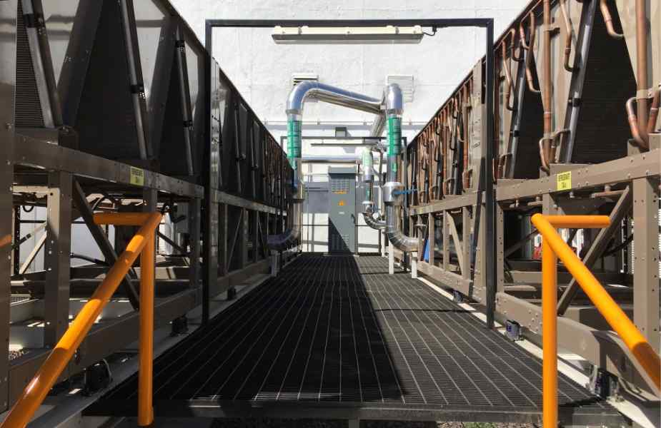 A narrow pathway flanked by large cooling units leads to a gray control door, with orange railings and industrial piping visible.