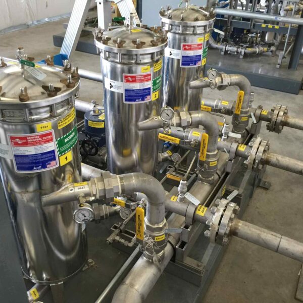Industrial filtration system featuring stainless steel filters, valves, and pipes in a well-lit facility with safety labels.