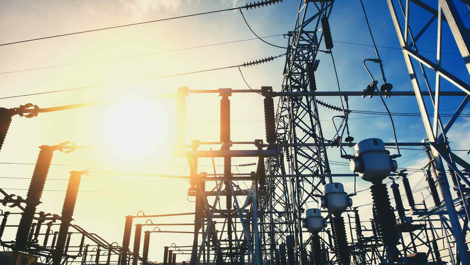 Silhouette of power lines and electrical infrastructure against a bright sunset sky, highlighting the energy distribution network.