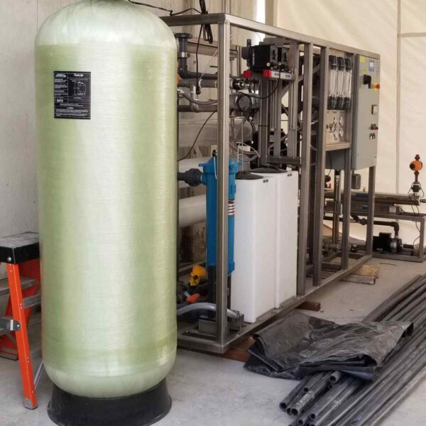 A green pressure tank beside a metal frame with various pipes and a control panel, set in a storage area with black plastic and metal pipes.