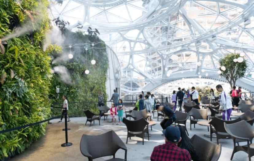 A bustling indoor garden space with lush greenery, misting features, and visitors relaxing in modern chairs beneath a glass dome.